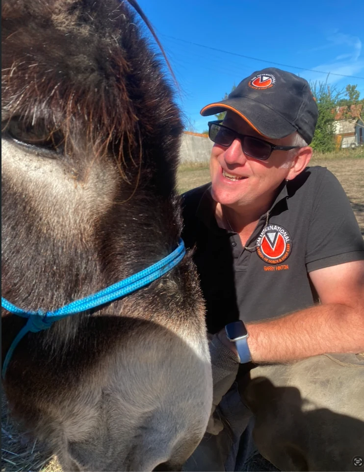 Garry Hinton, in an HM cap and polo shirt, smiling beside a donkey wearing a blue headcollar